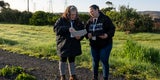 Two people stand on a path near green grass and other vegetation looking at papers.