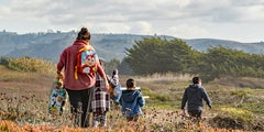 Woman in red jacket with childs backpack walks behind three children on coastal bluff