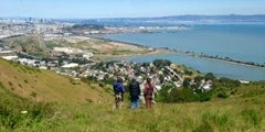 aerial view of mountains, water, the city with three people standing on the mountain looking towards the city