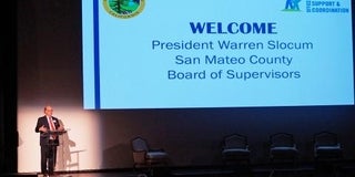 Presenter standing next to a welcome slide projected on screen