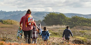 Woman in red jacket with childs backpack walks behind three children on coastal bluff