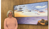 Artist Christine stands in a pink sweater next to her painting "Journey's End" which features a purple sea and golden sunset and hiking boots on a rock
