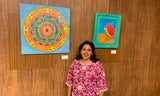 Woman in bright pink shirt stands with 2 colorful paintings