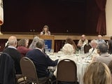 Jackie behind podium speaking to people sitting at round tables