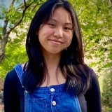 Teen youth with dark shoulder-length hair, smiling face forward. She is wearing a dark blouse with jean overalls.