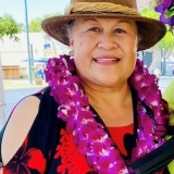 Woman with a hat on with her hair pulled back and she is smiling face forward. She is wearing a red and black blouse and a purple flower lei.