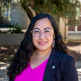Supervisor Noelia Corzo is a Latina woman with long, curly black hair and glasses. She is wearing a dark suit with a magenta blouse.