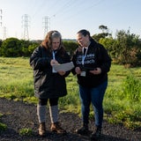 Two people stand on a path near green grass and other vegetation looking at papers.