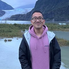 smiling man with glasses wearing a lavender hoodie sweatshirt and blue jacket. Background is a lake and glacier