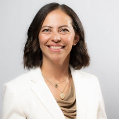 woman with brown hair and light eyes wearing a cream jacket and tan shirt